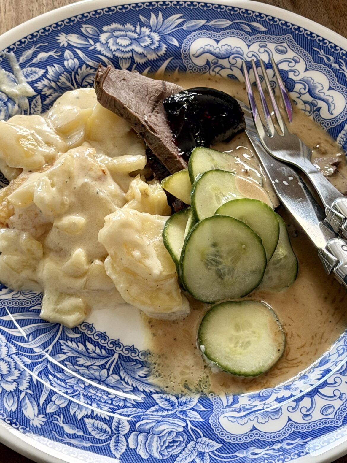 Krämig potatisgratäng serverad med skivat kött, gelé, pressgurka och gräddsås på en blåvit porslinstallrik.